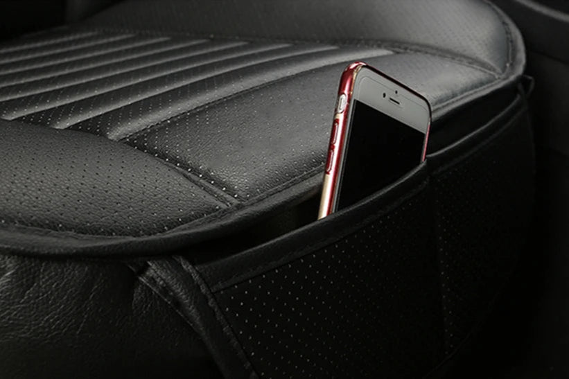 angled view of a black backless car seat cushion; perforated leatherette surface and a slim side pocket with a phone partially inserted