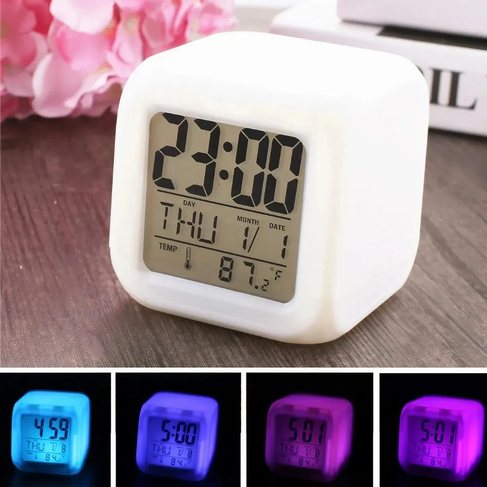 Digital LED alarm clock with a monochrome display set to 23:00, placed on a wooden desk near pink flowers; bottom row shows the clock lit in various colors including blue, purple, and pink.