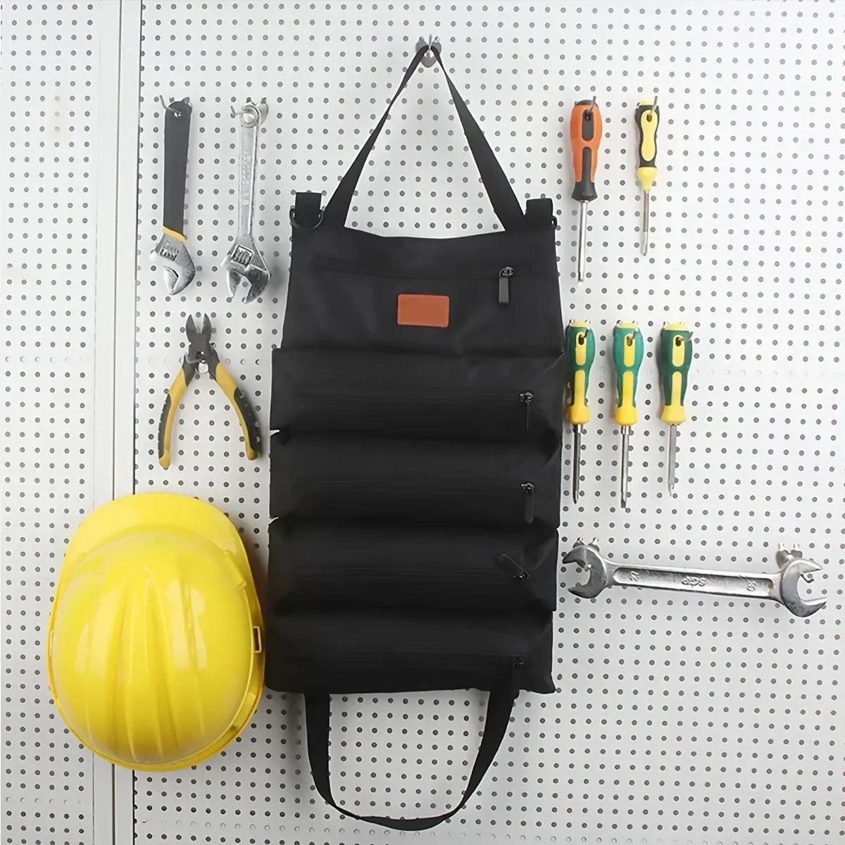 Black canvas tool roll bag hanging on a pegboard wall surrounded by various hand tools and a yellow hard hat, showcasing organized storage for workshop or garage settings.