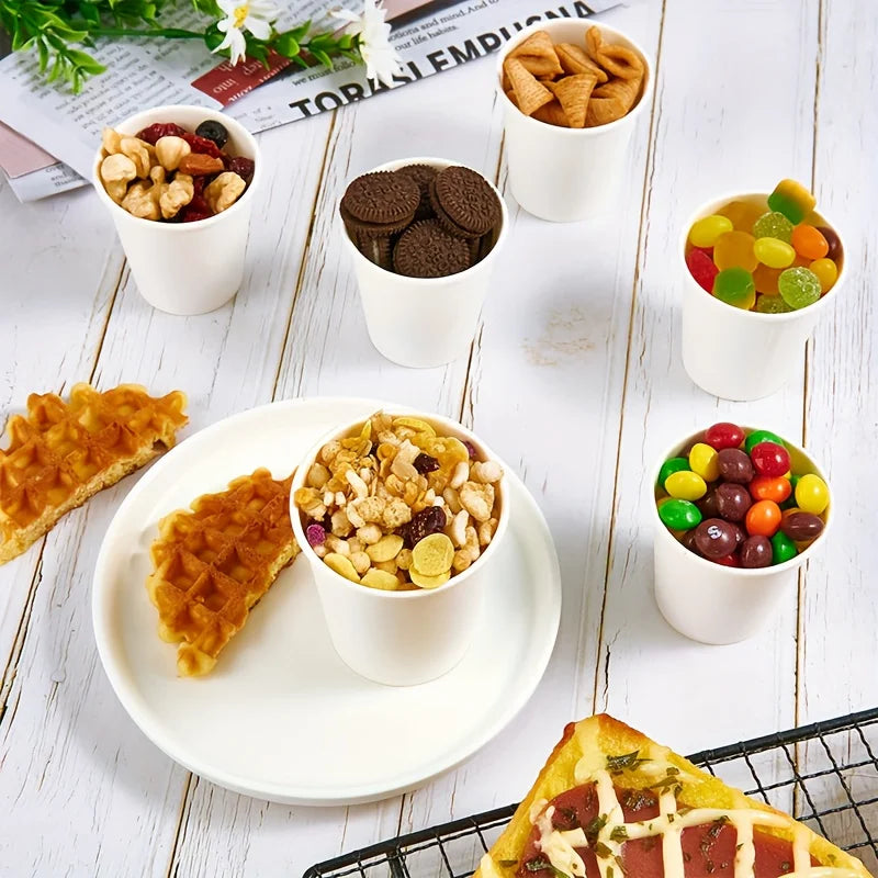 Small white paper cups filled with candy, cookies, and snacks on a white wooden table.