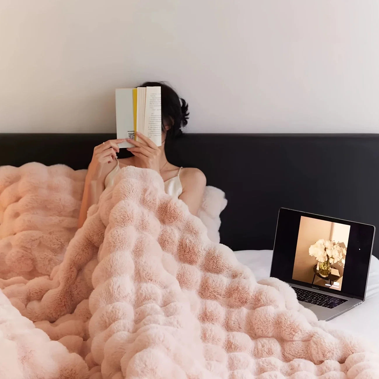 Person reading in bed under a blush pink faux-rabbit fleece blanket, laptop on the duvet, soft minimalist bedroom.