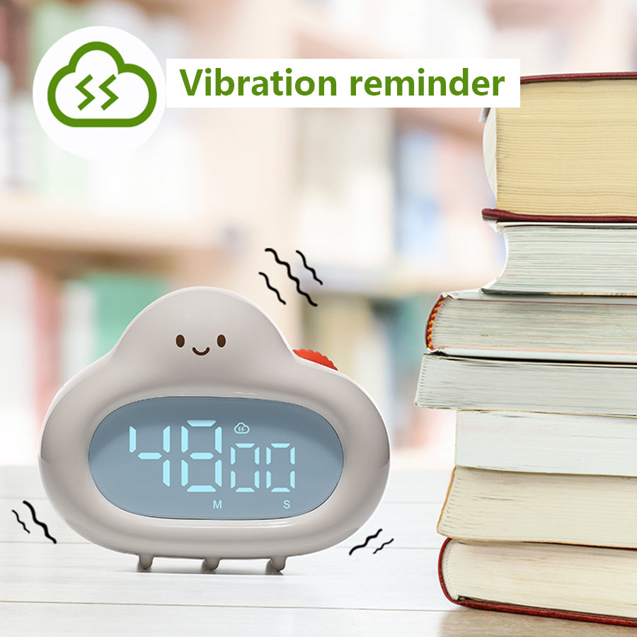 A white cloud-shaped timer placed on a table beside a stack of books, with digital numbers reading “42:00” on its blue-lit screen. Wavy lines around the timer suggest vibration. A green icon and the label “Vibration reminder” appear in the top left corner, indicating the vibration alert feature.