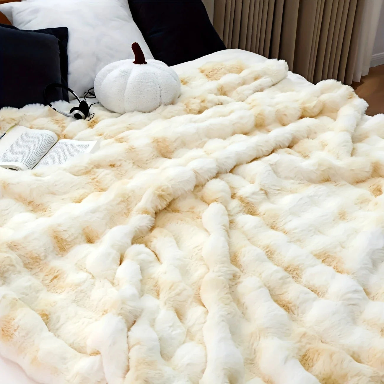 Cream faux-rabbit fleece blanket laid across a bed with headphones and an open book.