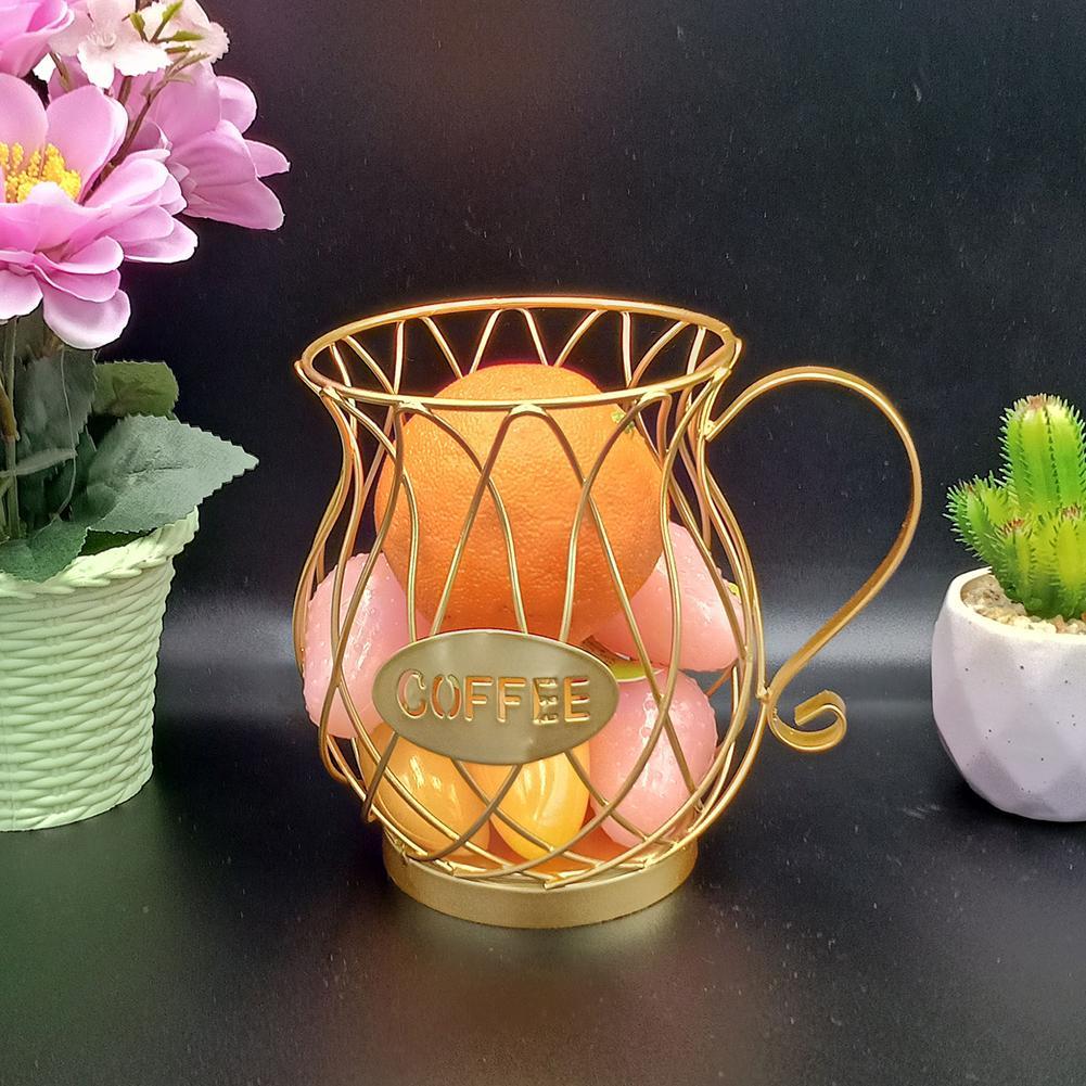 A gold coffee capsule basket repurposed to hold oranges and pink artificial fruits, surrounded by potted flowers and succulents on a dark black background, emphasizing decorative versatility.