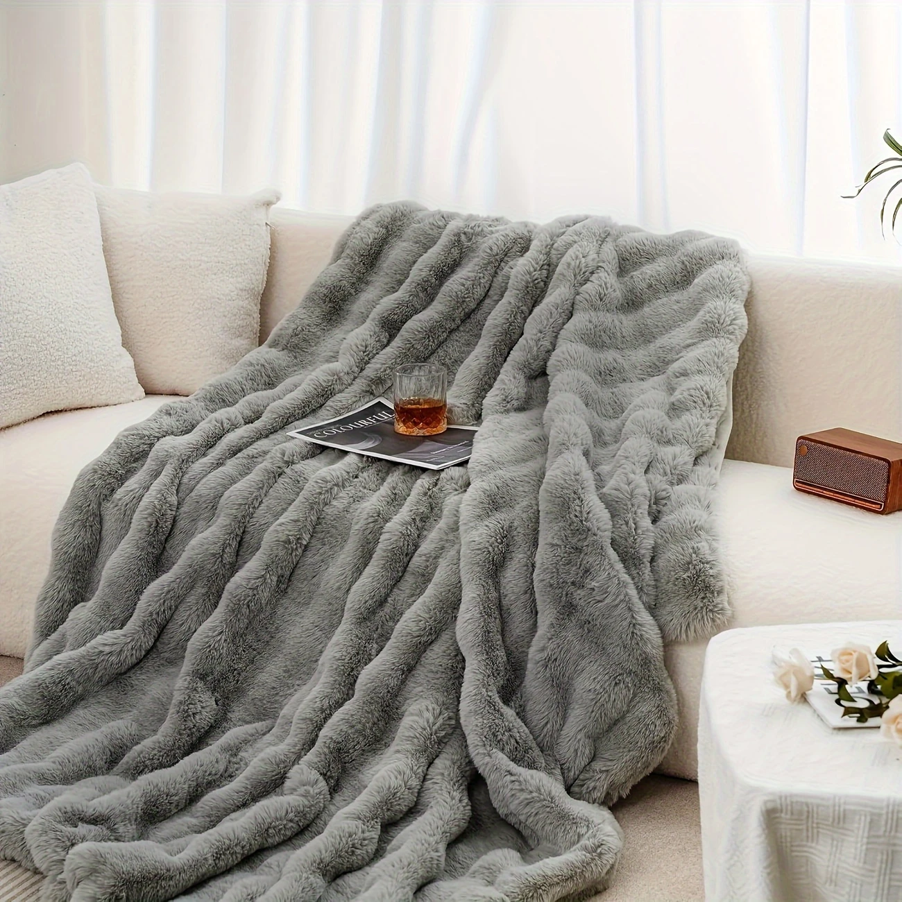 Light gray faux-rabbit fleece blanket flowing over a sofa arm with a magazine and drink on the side table.