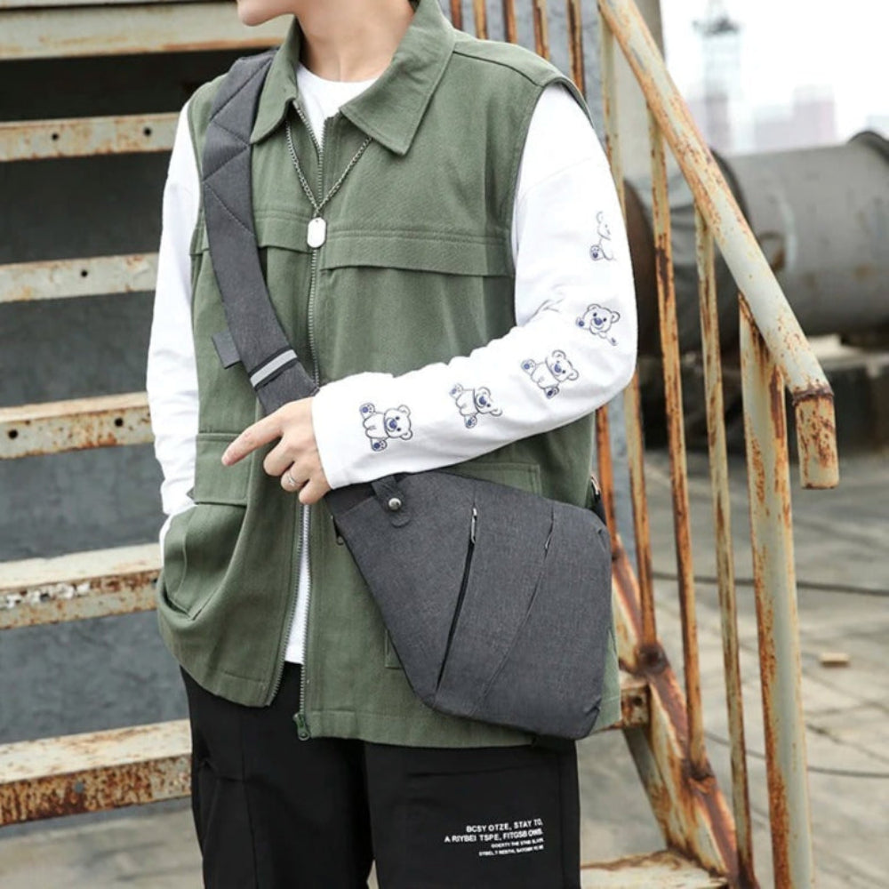 Male Model with streetwear on wearing a gray crossbody bag next standing next to a rusted stair case. 