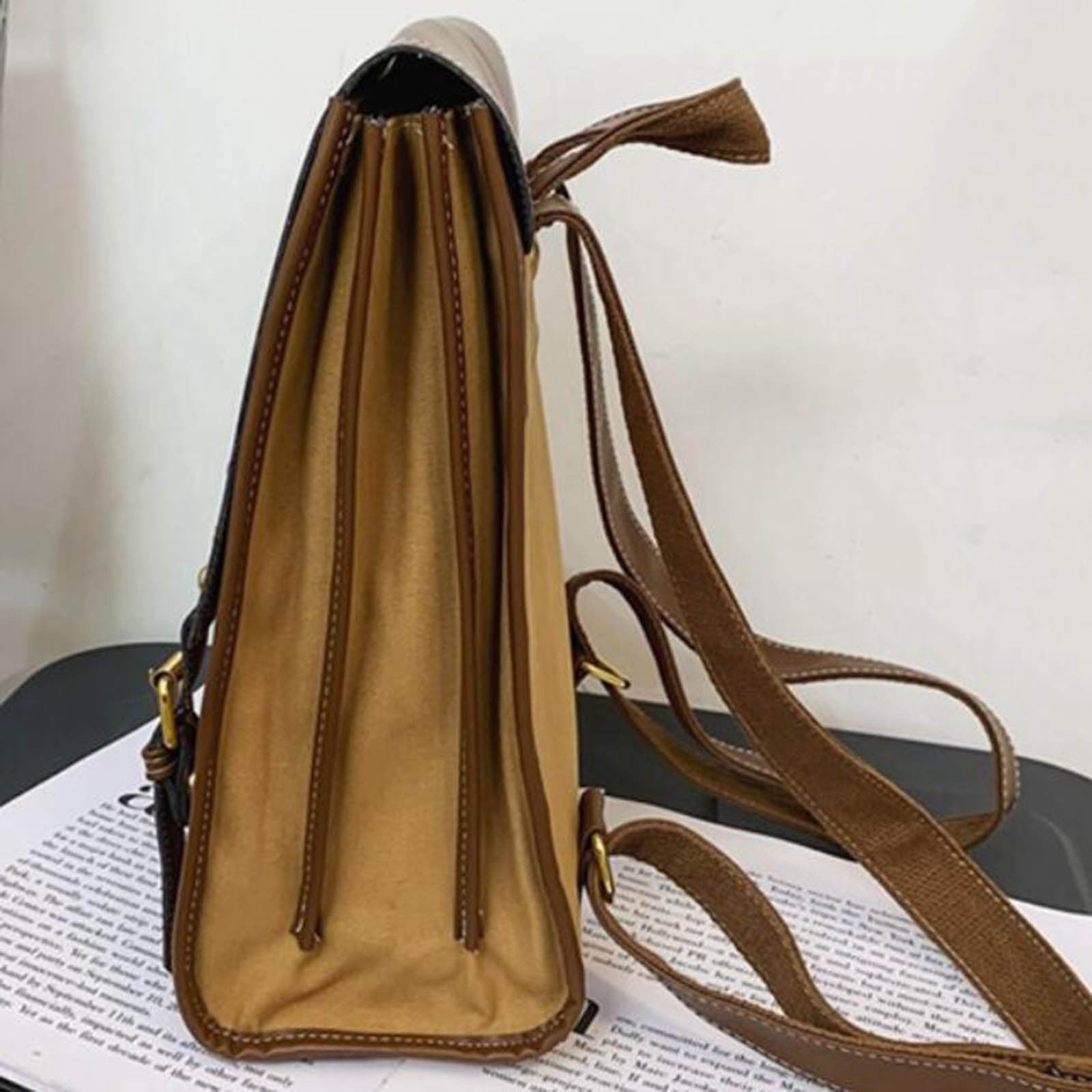 Side view of the backpack showing brown canvas fabric with expanding pleats and adjustable shoulder straps resting on a surface with text.