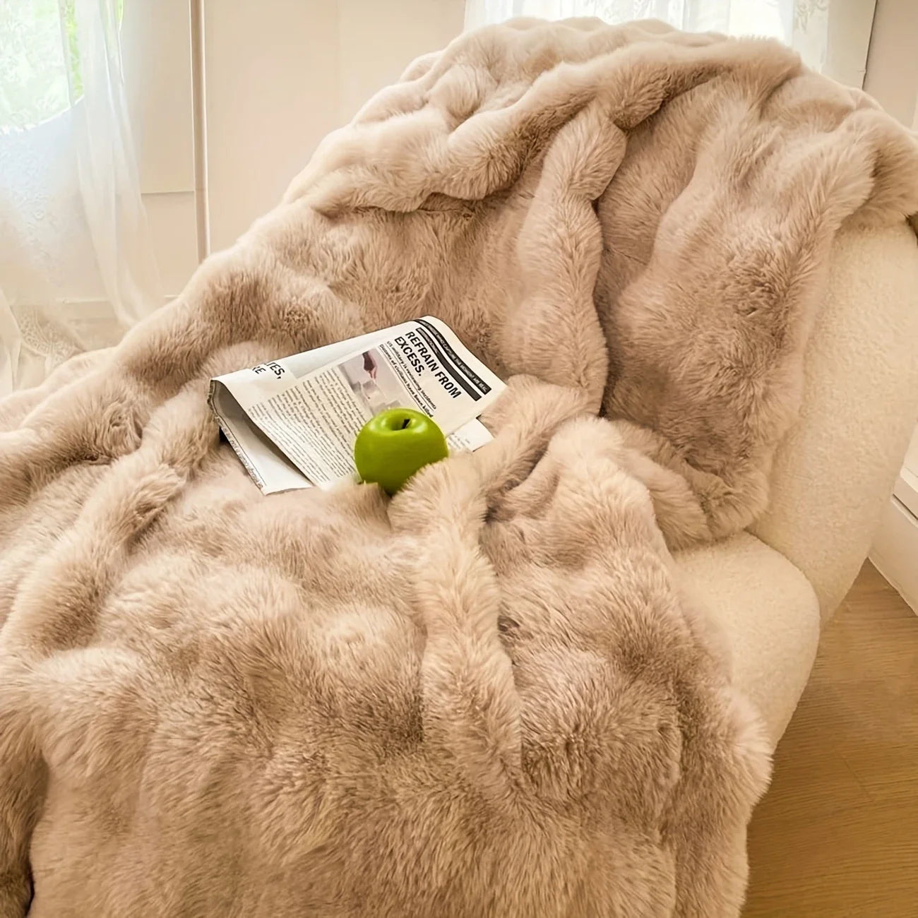 Taupe faux-rabbit fleece blanket spread on a bouclé chair with a newspaper and green apple.