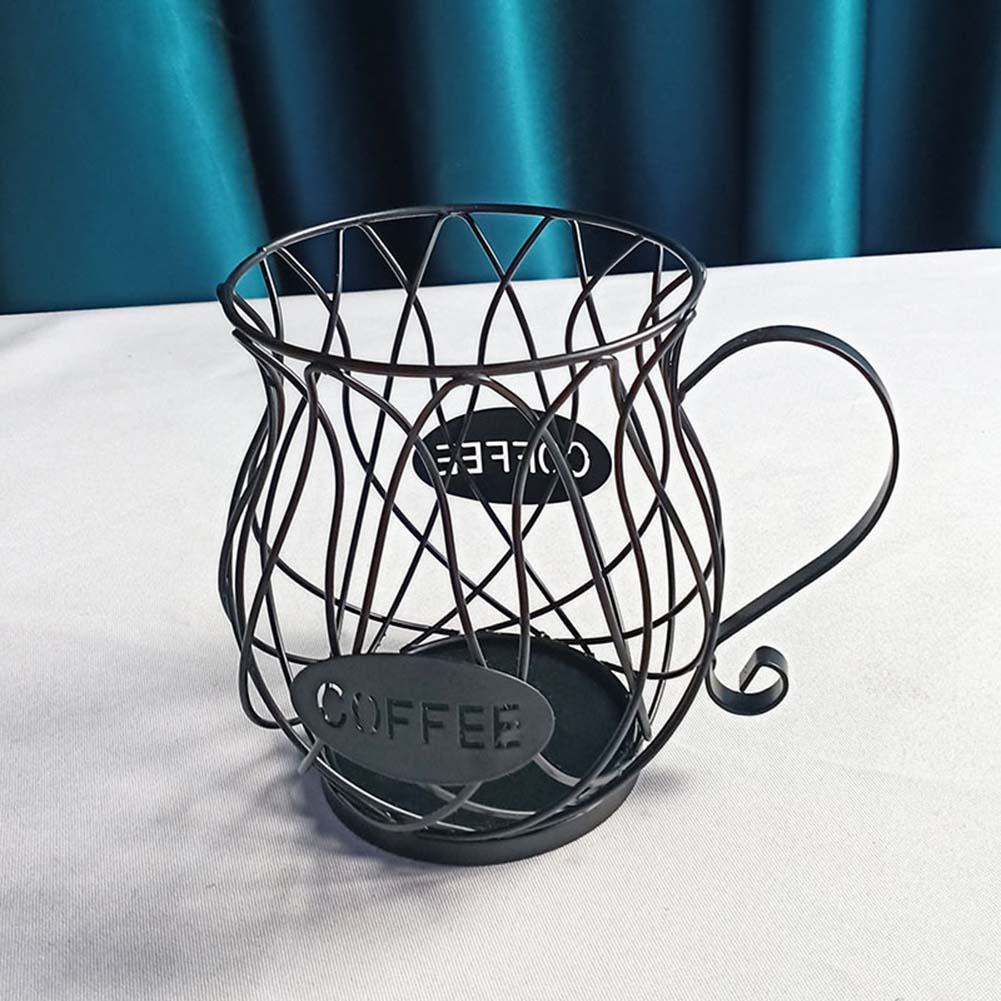 A matte black iron wire coffee capsule holder in a cup shape, placed on a white table with teal drapery in the background, showcasing its elegant lattice structure and labeled "COFFEE."