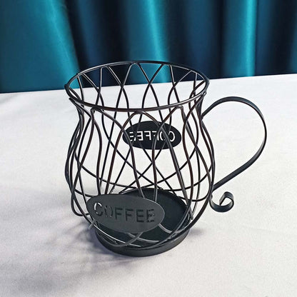 A matte black iron wire coffee capsule holder in a cup shape, placed on a white table with teal drapery in the background, showcasing its elegant lattice structure and labeled "COFFEE."