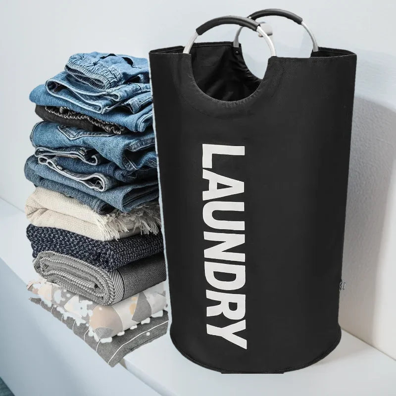 A black cylindrical laundry hamper with aluminum handles and the word "LAUNDRY" printed in white, shown beside a neatly stacked pile of folded jeans, blankets, and towels on a laundry counter in a modern home.