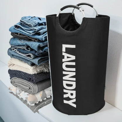 A black cylindrical laundry hamper with aluminum handles and the word "LAUNDRY" printed in white, shown beside a neatly stacked pile of folded jeans, blankets, and towels on a laundry counter in a modern home.