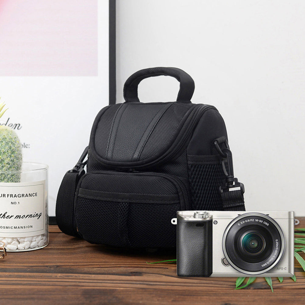 Black camera shoulder bag displayed on wooden surface beside a mirrorless camera, featuring front mesh pocket and padded top handle.