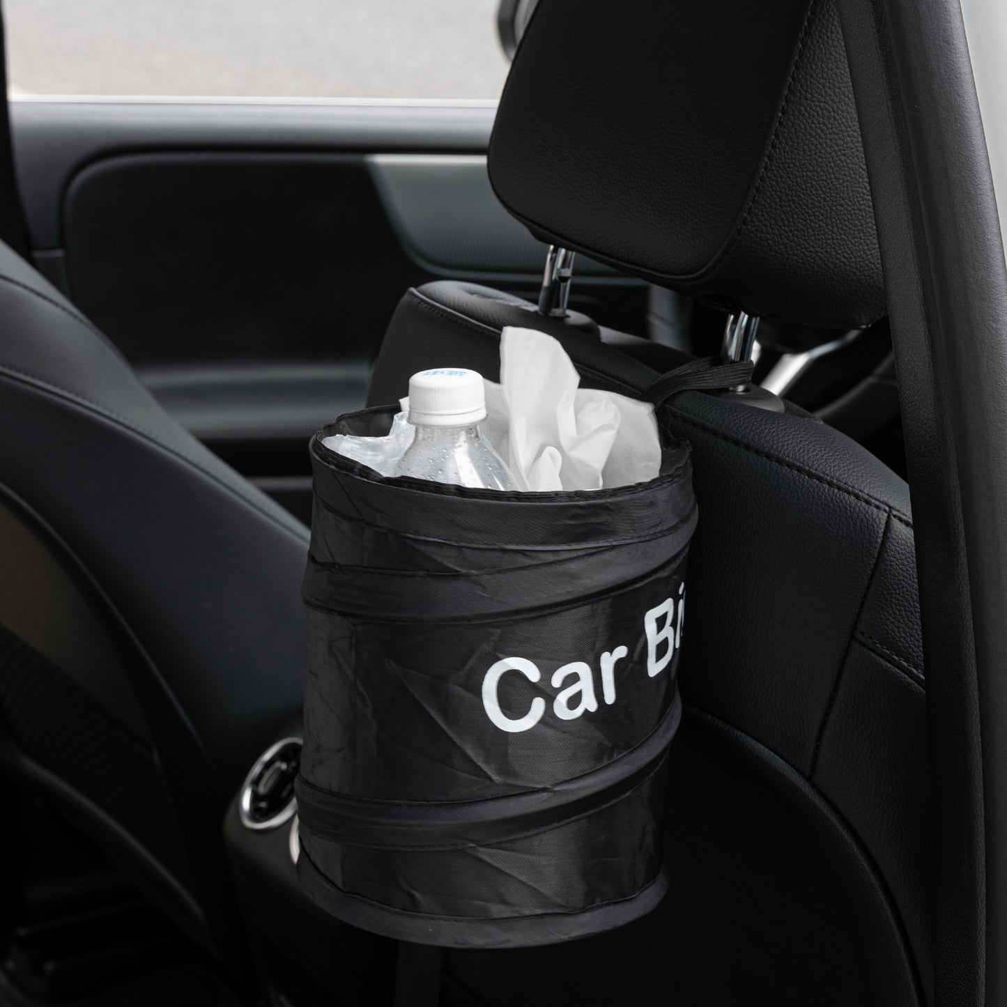 Portable car bin positioned beside the front console of a luxury vehicle, holding drink bottles and tissues, with caption “It holds the trash in place.