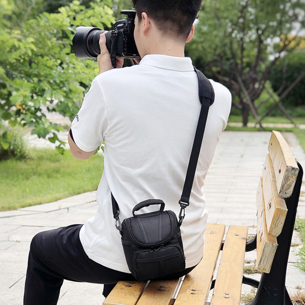 A photographer wearing a compact black shoulder camera bag while using a DSLR camera outdoors. The bag is lightweight and designed for convenient travel and everyday use.