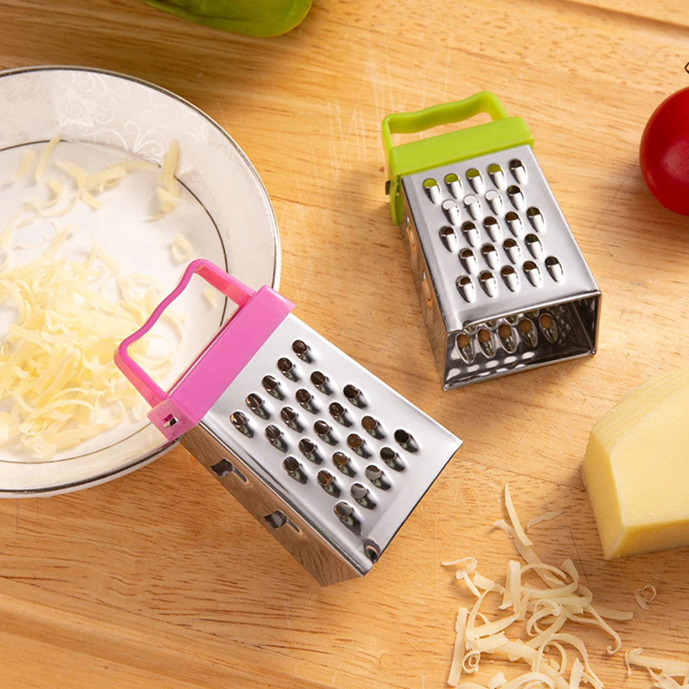 Pink and green mini stainless steel box graters on wooden surface beside shredded cheese and tomatoes