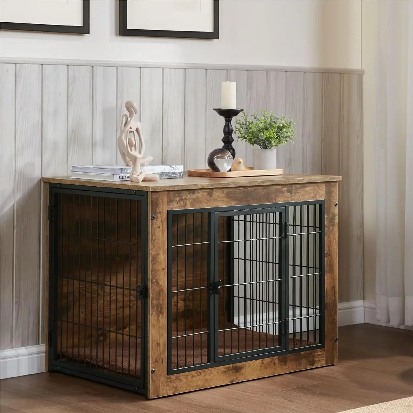 Rustic indoor dog crate displayed as functional furniture in a bright room. Features a wooden top surface with a potted plant, candle, sculpture, and book decor, showcasing its use as a side table. The crate’s front panel includes a wide-opening black metal door and a visible soft brown mat inside.